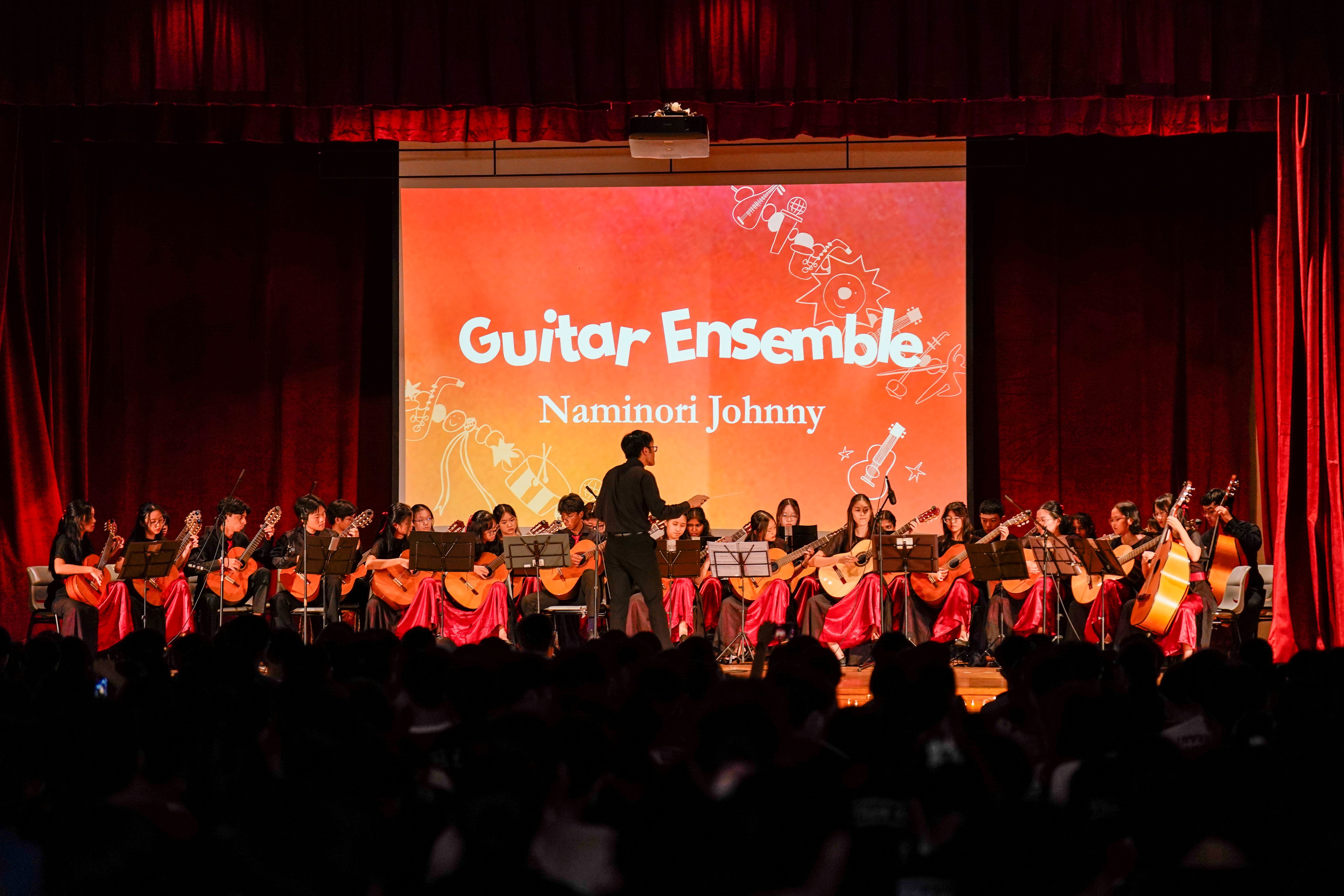 A Guitar Ensemble performs "Naminori Johnny" on stage, with students seated in a semicircle holding guitars while a conductor leads them.
